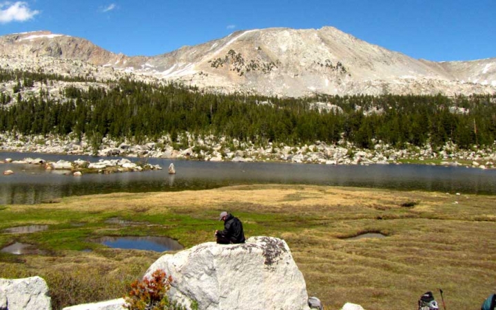 A person sits on a tall rock above a grassy landscape and appears to journal. There is a body of water, evergreen trees and mountains in the background.